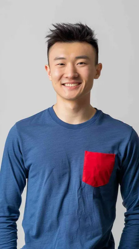 A young Chinese man with short black hair, wearing a blue long-sleeved shirt with a red pocket on the chest area of his right hand is smiling at the camera. The background color should be white to highlight his face. He has an Asian nose shape, medium skin tone, round eyes, and thin eyebrows. His overall appearance conveys friendliness or confidence. in the style of --ar 25:32 - style2部分