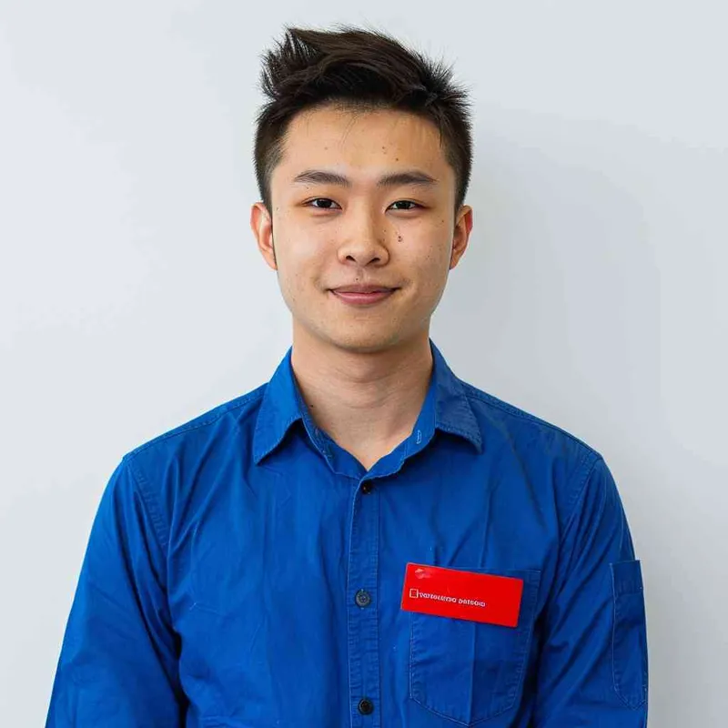 A young Chinese man with short black hair, wearing a blue long-sleeved buttoned shirt and a red name tag on his chest, smiling at the camera against a white background for a professional profile picture. The image is in the style of a typical company profile photo. --ar 25:32 - style3部分